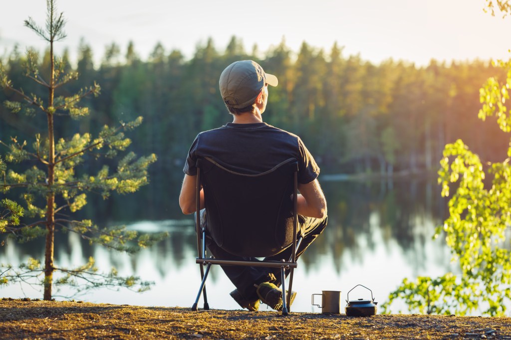 Un hombre se sienta en una silla de camping con el telón de fondo de un lago forestal en una tarde de verano.