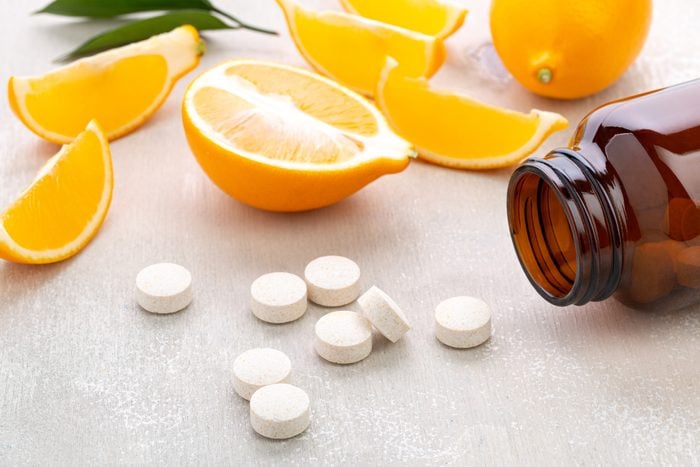 Las vitaminas se encuentran cerca de frascos de vidrio oscuro con rodajas de limón y frutas enteras sobre una mesa de madera blanca.