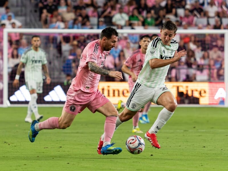 Dos jugadores de fútbol, uno con una camiseta rosa y el otro con una camiseta blanca y verde, persiguen el balón durante un partido, con la multitud y otros jugadores borrosos en el césped al fondo.