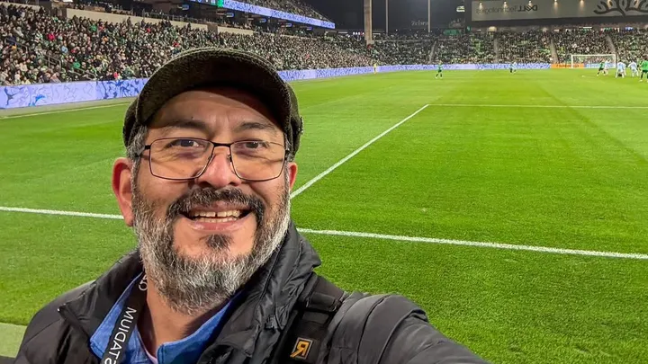 Un hombre con gafas, sombrero y chaqueta sonríe y se hace un selfie en un estadio de fútbol, con un campo verde y gradas repletas de espectadores al fondo.