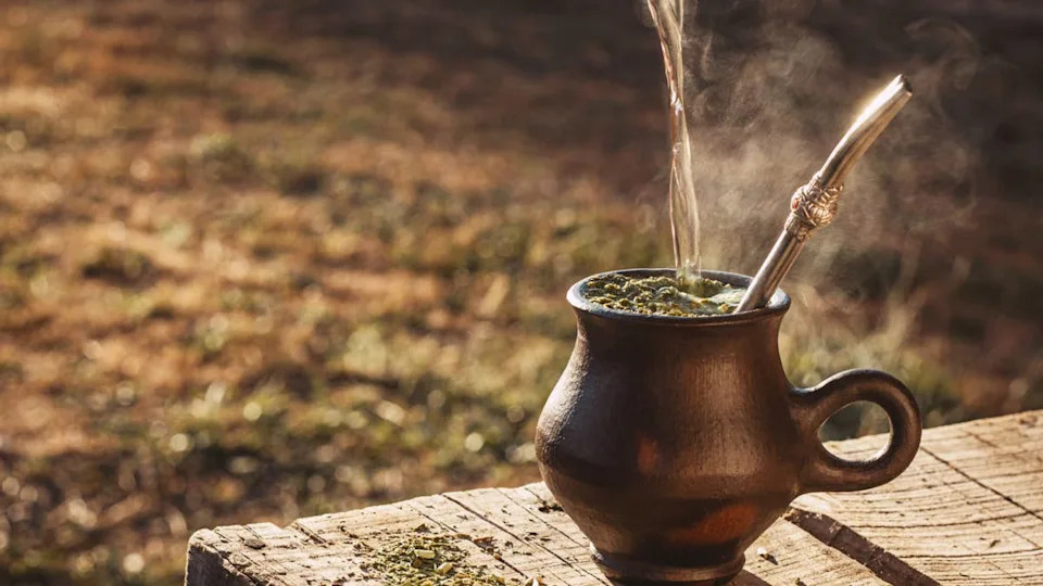Se sirve agua en tazas humeantes de yerba mate en una mesa rústica bajo luces doradas, sobre un fondo de hierba verde. Sebastián Soto/Getty Images